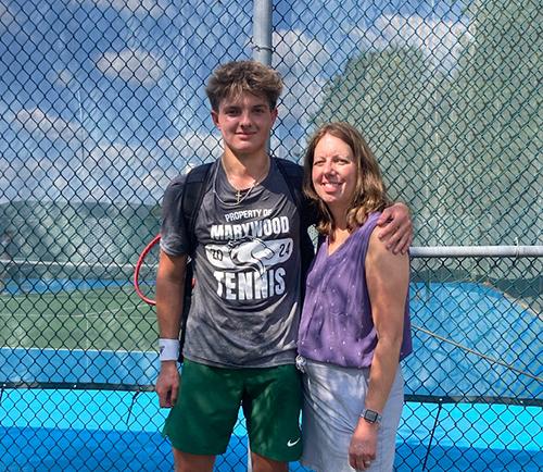 boy in grey marywood tennis shirt and green shorts standing in front of tennis court next to middle aged woman in purple tank top with his arm around her shoulder